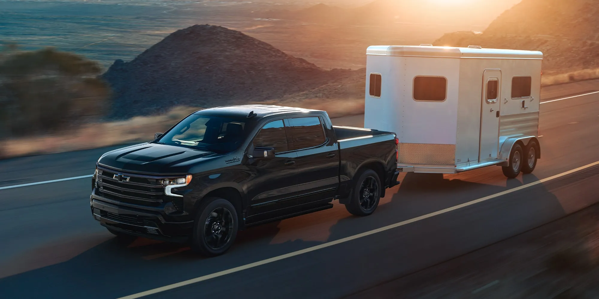 A black Chevrolet Silverado towing a horse trailer on a highway at sunset