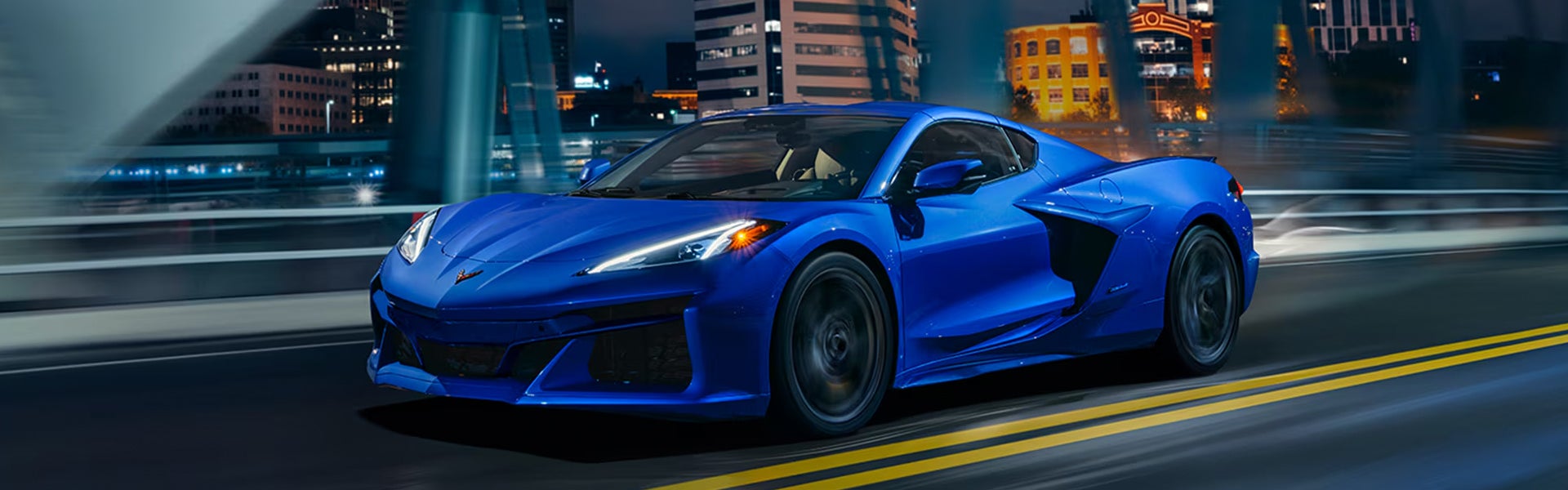 2026 Chevrolet Corvette E-Ray in bright blue driving across a city bridge at night, highlighting its sleek mid-engine design, aerodynamic body, LED headlights, and electrified high-performance sports car styling.