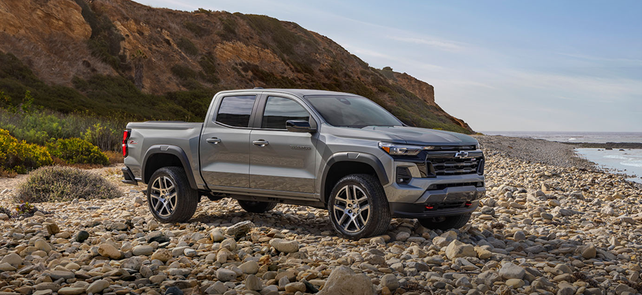 A silver 2025 Chevrolet Colorado is parked on a rocky beach with cliffs and greenery in the background.