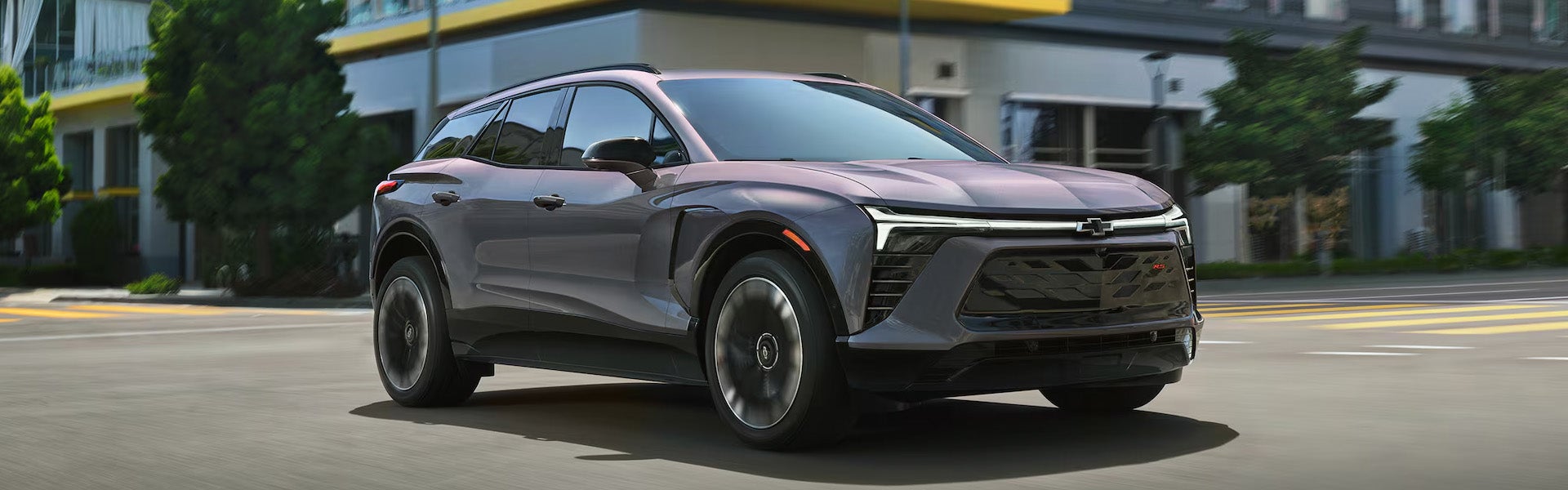 2026 Chevrolet Blazer EV in gray driving through a city street, showcasing its sleek aerodynamic design, distinctive LED lighting, bold front grille, and modern electric SUV styling.