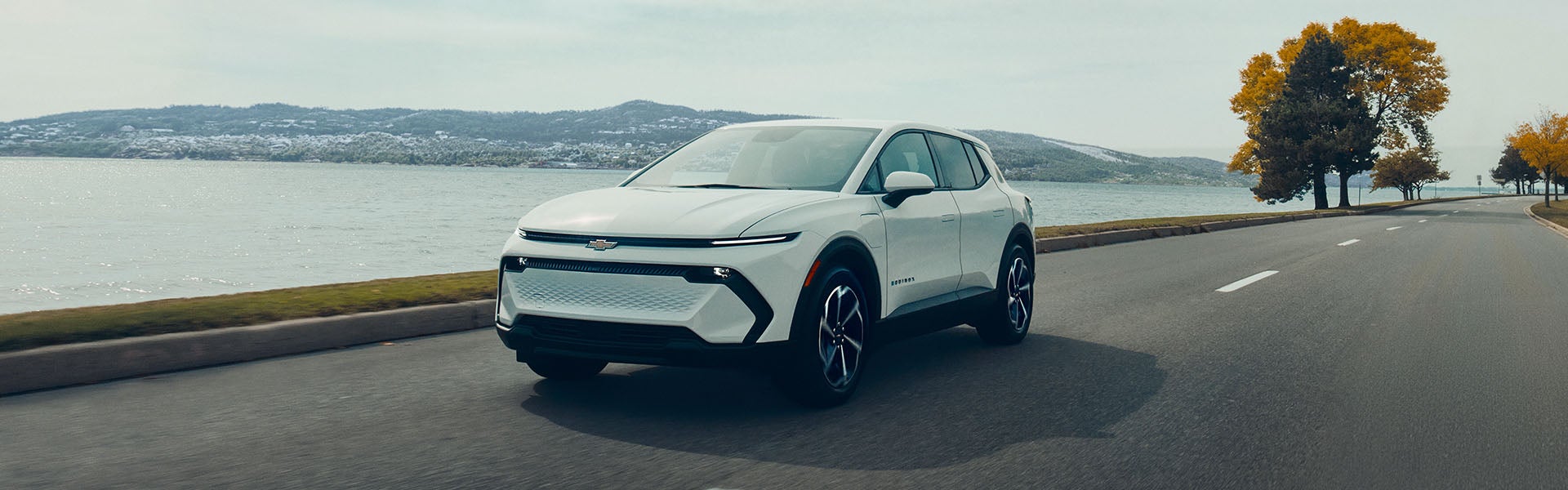 2026 Chevrolet Equinox EV driving along a lakeside road with green trees in the background.