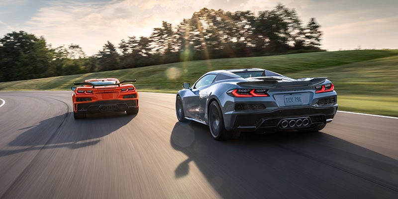 Two sports cars Chevrolet-Corvette-Z06, one silver and one orange, speed along a curving track with lush greenery and a sunlit sky in the background, conveying excitement and motion.