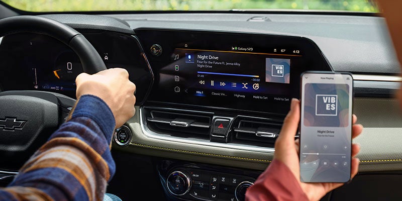 Inside a Chevrolet Trailblazer, a person drives while holding a smartphone displaying music synced to the car's touchscreen, showing "Night Drive."