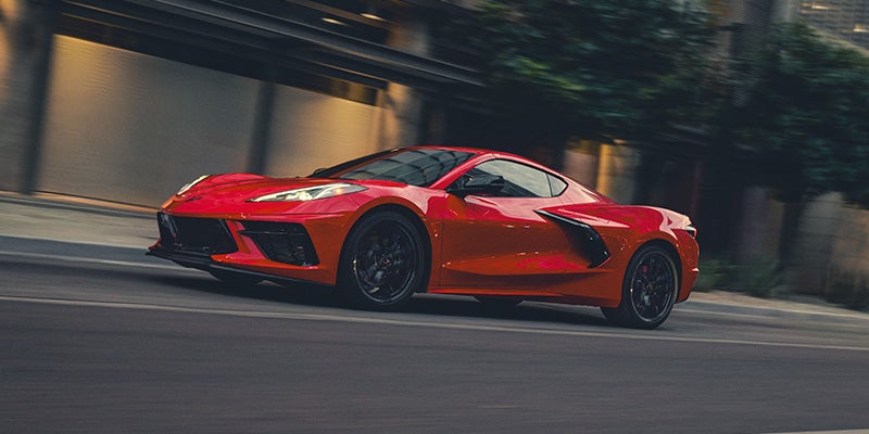 A sleek red sports Corvette Stingray speeds down a city street, conveying a sense of motion and excitement. The blurred background enhances its dynamic movement.