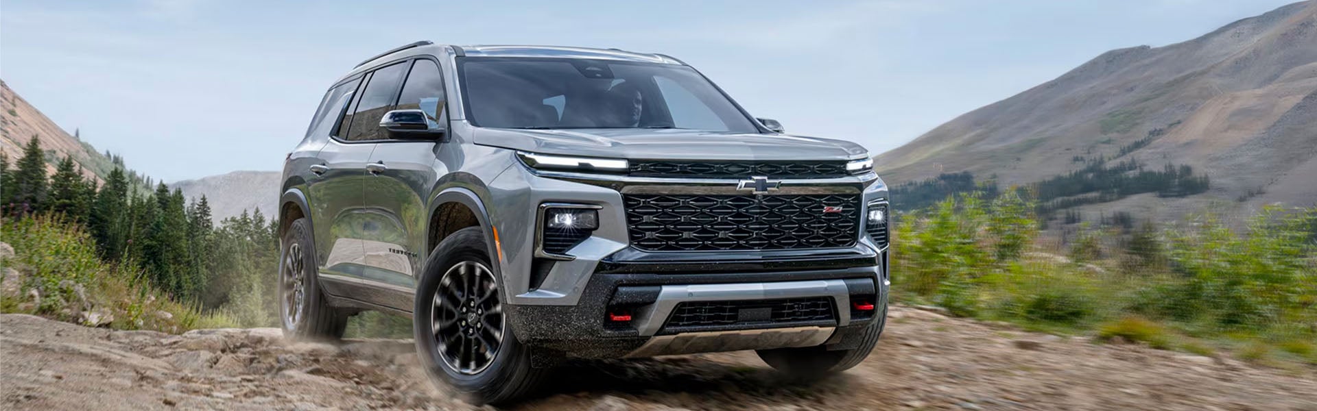 2026 Chevrolet Traverse in Silver driving on a rugged dirt road with mountainous scenery. SUV bold design and dynamic stance convey adventure and power.