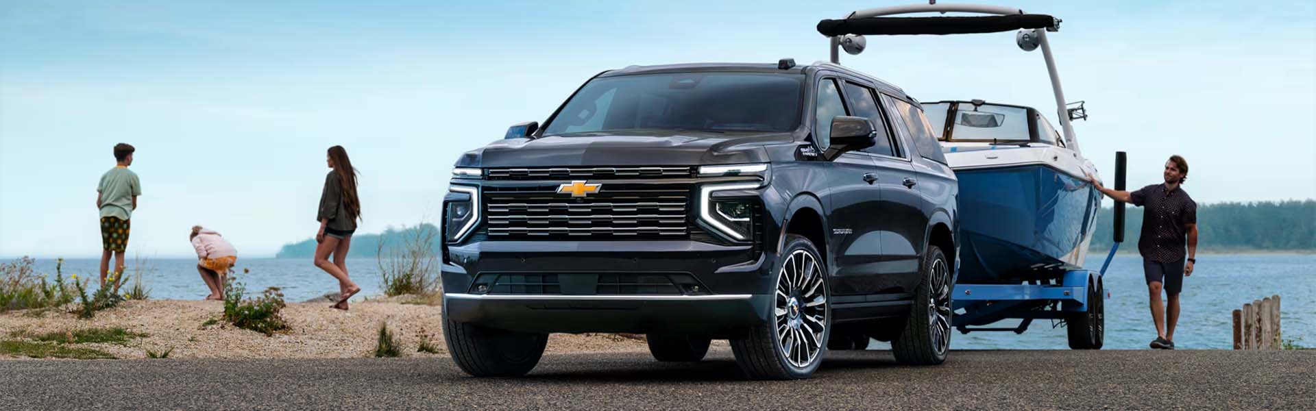 A black 2025 Chevrolet Suburban towing a blue recreational boat at a lakeside launch area.