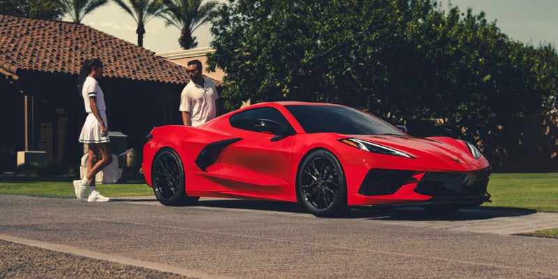 A sleek red sports Corvette Stingray is parked in front of a house with a tiled roof and palm trees. Two people stand nearby, conveying a lifestyle of luxury and leisure.