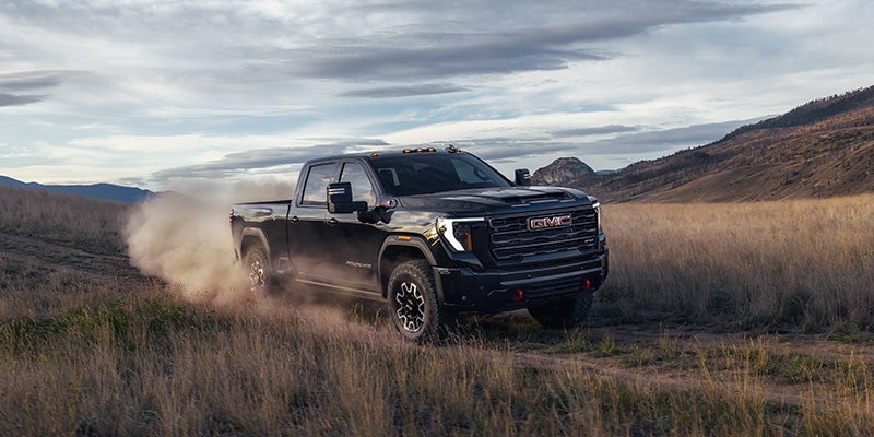 A black pickup GMC Sierra HD kicks up dust while driving through a grassy, rugged landscape under a cloudy sky, conveying power and adventure in a natural setting.