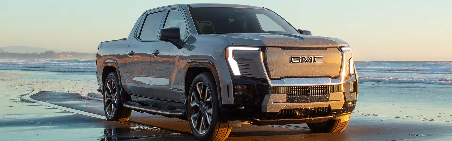 A silver 2026 GMC Sierra EV parked on a wet beach with waves in the background.