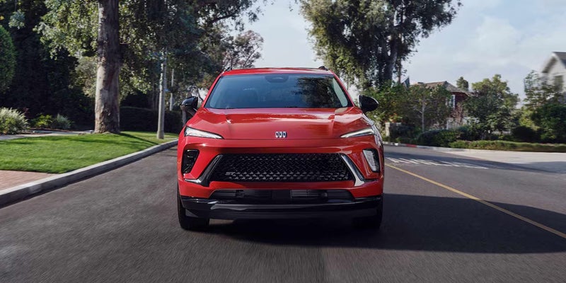 A red Buick Envision drives down a suburban road lined with trees and houses. The car's bold design and shiny finish enhance its modern, dynamic presence.