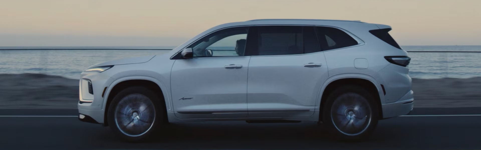 2026 Buick Enclave SUV driving along a coastal road at dusk, shown in side profile with the ocean in the background.