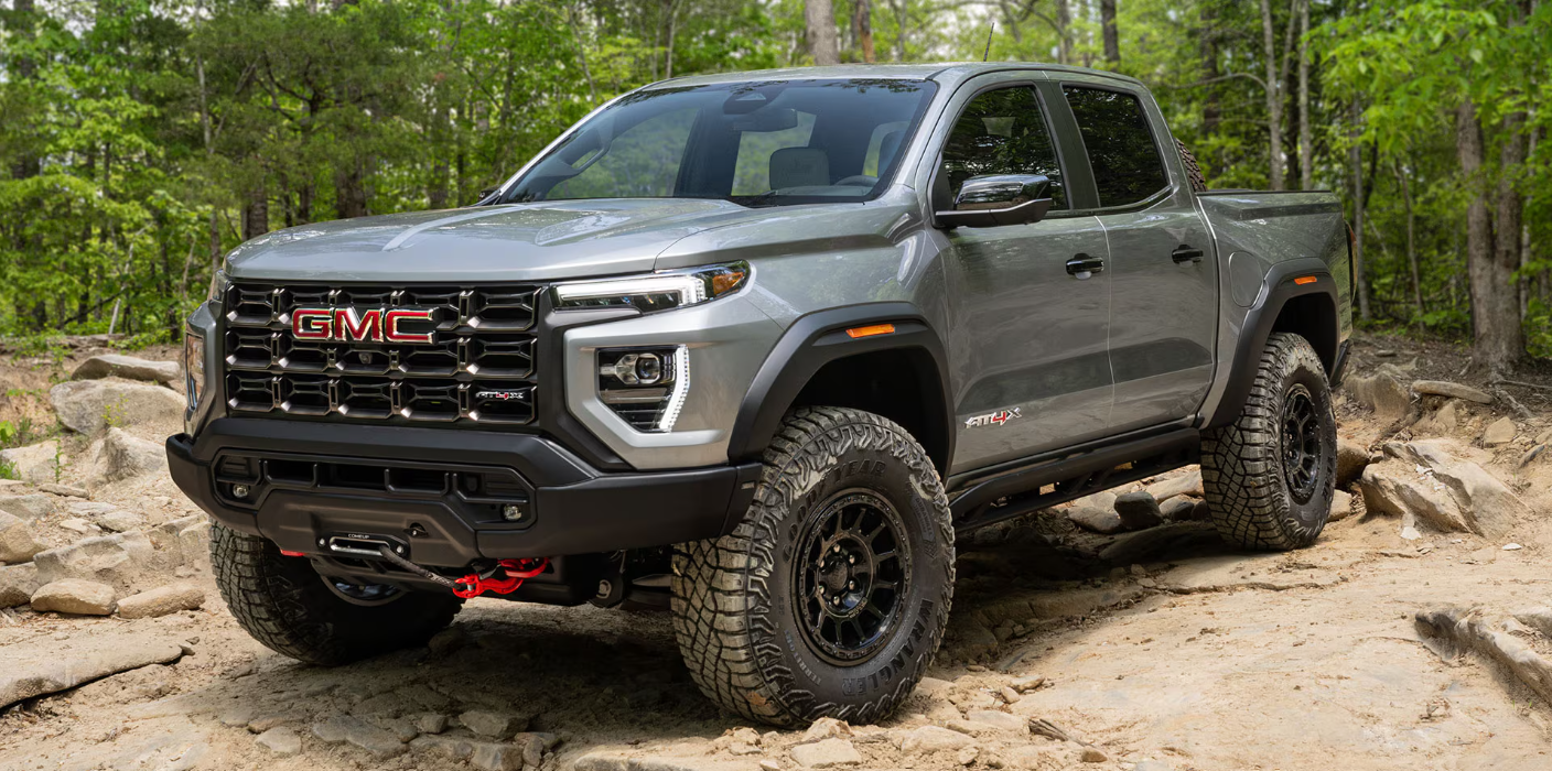 A rugged gray GMC AT4x pickup truck is parked on a rocky, forested path. The truck's design is bold with a prominent grille and large tires, conveying strength.