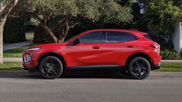 Red 2026 Buick Envision compact SUV parked on a quiet residential street with trees and greenery in the background.