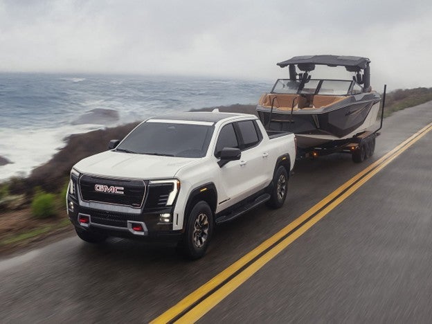 2026 GMC Sierra EV electric pickup truck pulling a boat trailer while driving on a scenic oceanfront road.
