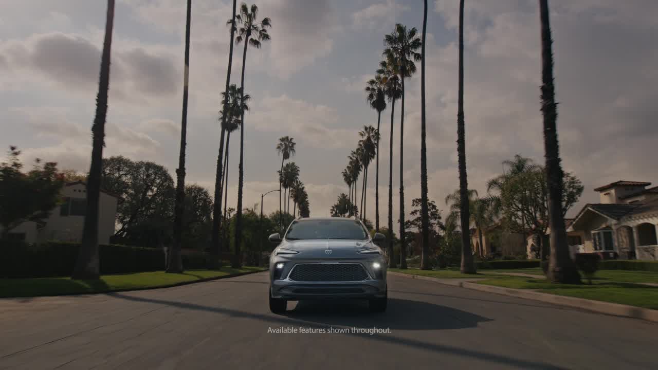 A 2026 Buick Encore GX Avenir drives down a street lined with palm trees, showcasing a sunny, tropical atmosphere.