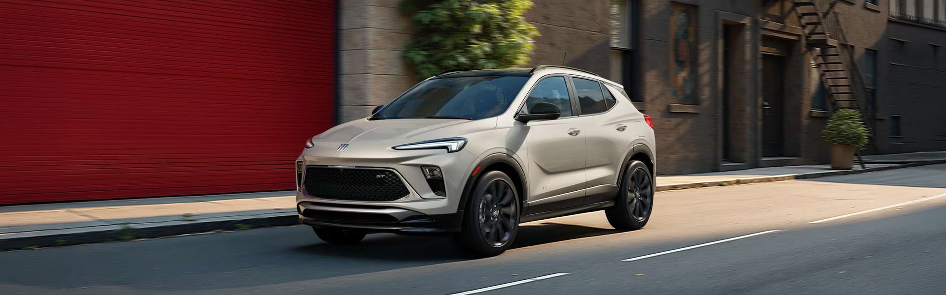 2026 Buick Encore GX driving past a red garage, showcasing its sleek design and sporty profile.