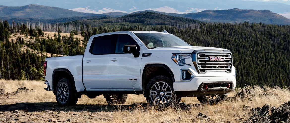 A white GMC pickup truck Sierra 1500 AT4 with rugged tires is parked on a grassy hill overlooking a vast forest and distant mountains under a clear blue sky.