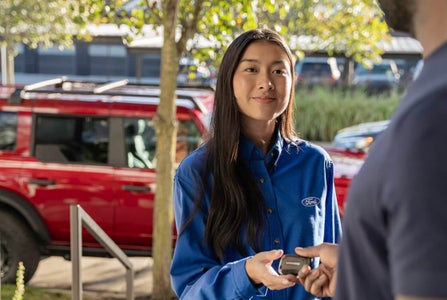 Close-up view of a Ford representative handing a vehicle key fob to a customer with a 2026 Ford Bronco Sport in Ruby Red Metallic.