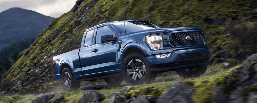 A blue pickup truck drives off-road on a lush, rocky hill. The backdrop features misty mountains and a cloudy sky, conveying adventure and ruggedness.