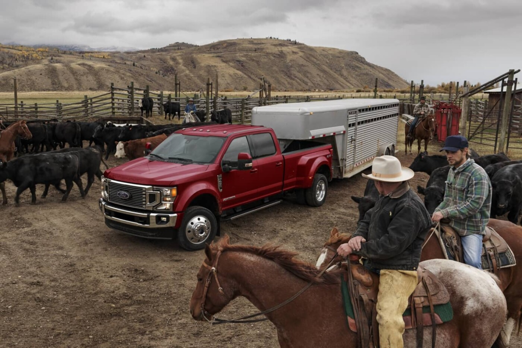 A red pickup truck with a cattle trailer is parked in a ranch setting. Two cowboys on horses guide cattle in a fenced area, with hills in the background.