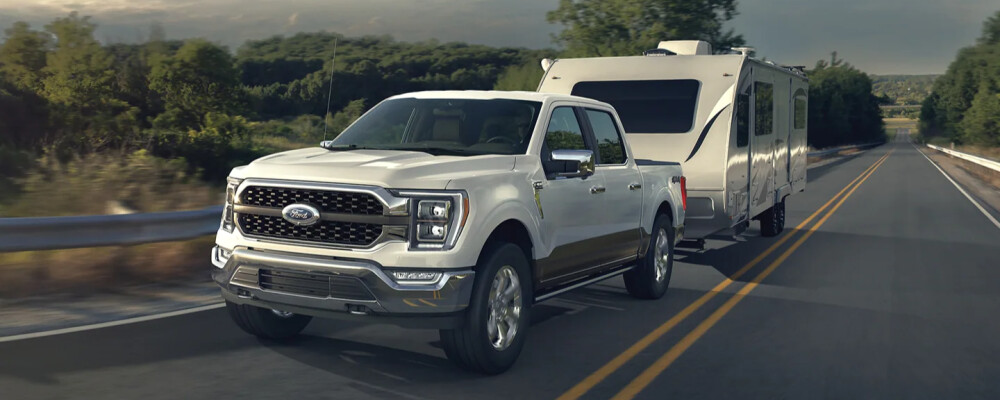 A white pickup truck towing a large RV trailer drives on a sunny rural road, surrounded by green trees. The scene conveys adventure and freedom.