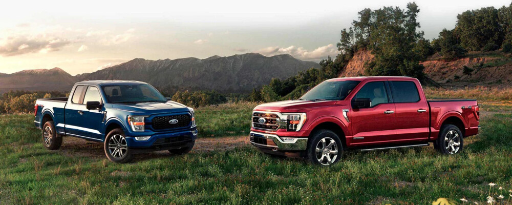 Two pickup trucks, one blue and one red, are parked on grassy terrain with a mountainous landscape in the background under a partly cloudy sky.