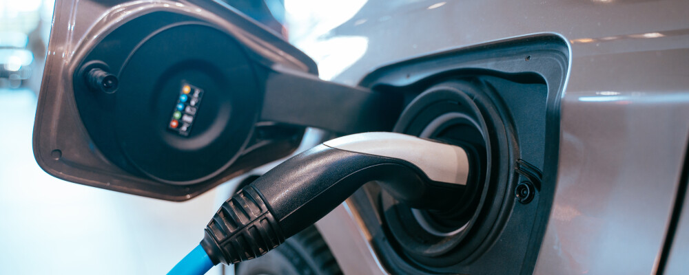 Close-up of an electric vehicle charging, highlighting a black and white plug connected to a gray car. The scene conveys modern, eco-friendly technology.