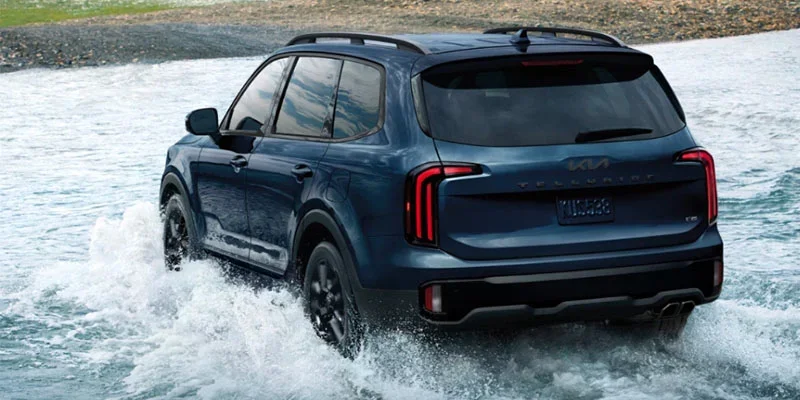 A blue Kia Telluride SUV driving through water, showcasing its rugged capability and sleek design. The vehicle's rear is highlighted as it makes its way through the water, with splashes surrounding it.