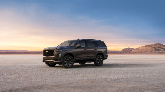 A sleek black SUV is parked on a vast, flat desert at sunset. The sky glows in shades of orange and blue, with distant rocky hills adding depth.