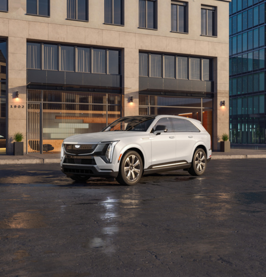 A sleek white SUV is parked on a wet city street in front of a modern building. The lighting suggests early evening, creating a polished, sophisticated ambiance.