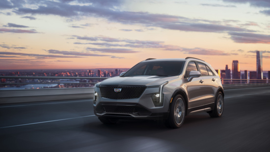 A silver cadillac xt4 SUV drives on a highway at dusk, with a city skyline and colorful sunset in the background, conveying a sense of elegance and adventure.