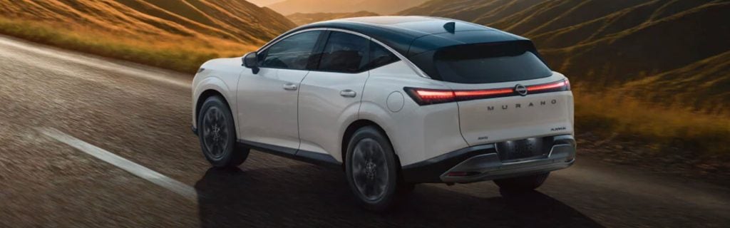 A white Nissan Murano driving on a paved road through a mountainous landscape during sunset, showing its rear three-quarter view.