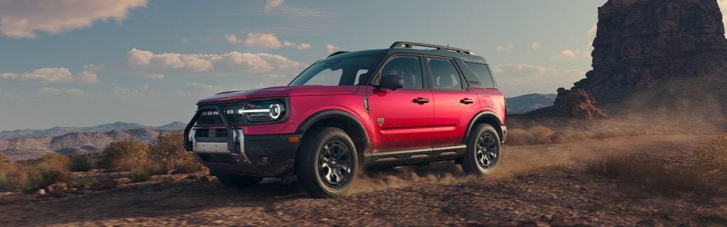 Exterior of a red 2025 Ford Bronco Sport suv, showing the front and driver's side, driving through rugged terrain. 