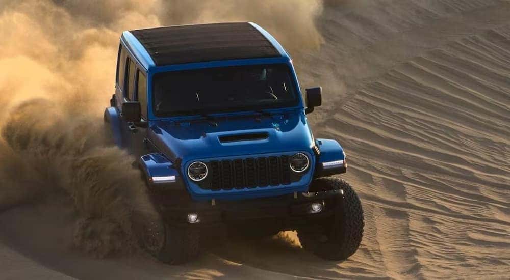 A blue 2024 Jeep Wrangler is shown off-roading on sand.