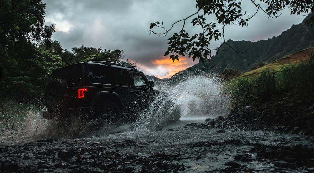 A black 2018 Jeep Wrangler Unlimited is driving through a river at dusk splashing up water.