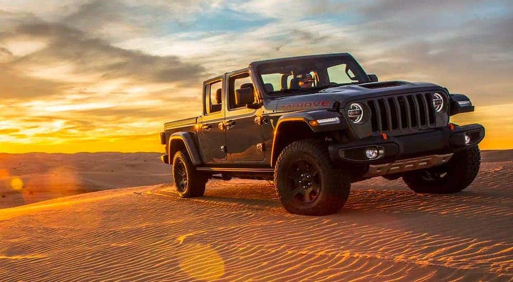 A gray 2020 used Jeep Gladiator Mojave with no roof is parked on sand dunes in front of a vibrant yellow sunset.