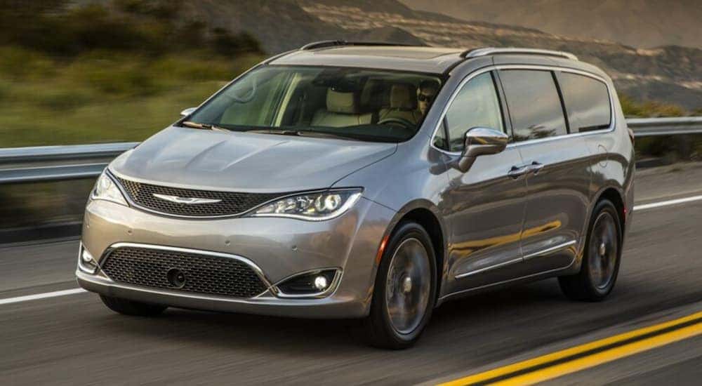 A silver 2017 Chrysler Pacifica is driving past a guard rail and mountains.