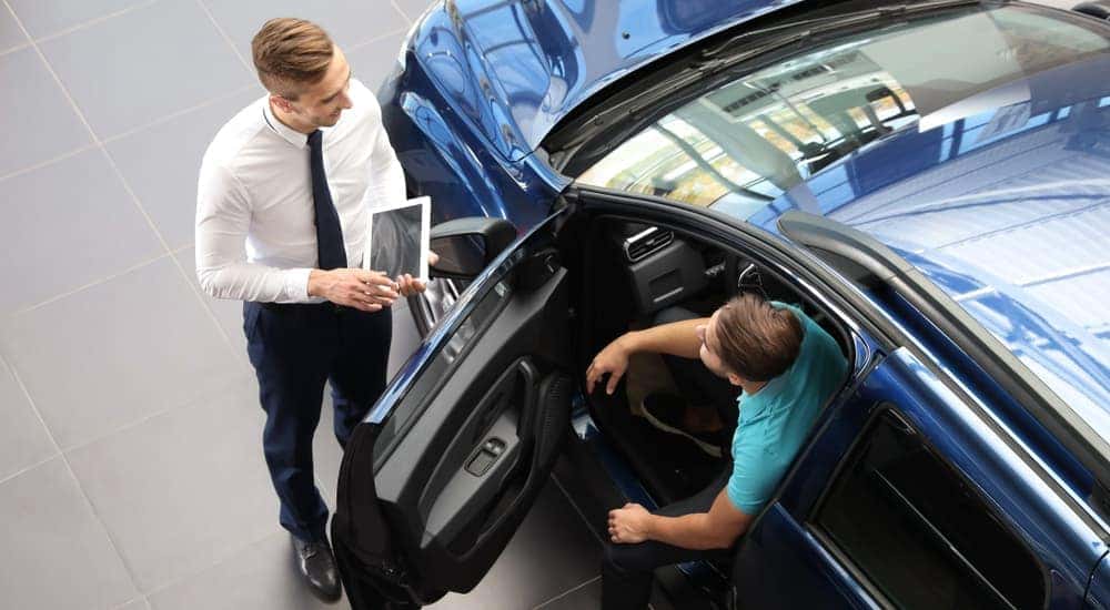 A customer is sitting in a blue car while talking to a salesman, shown from above.