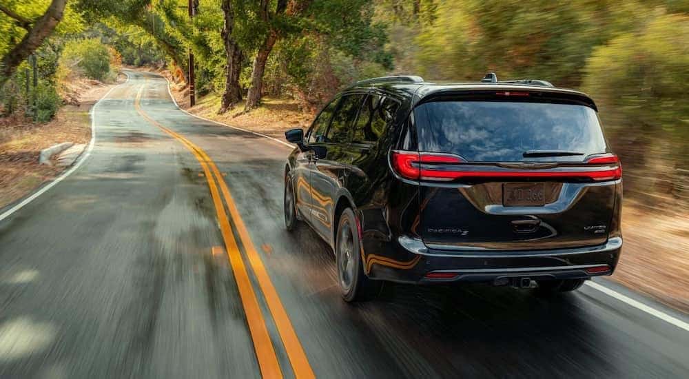 A black 2021 Chrysler Pacifica Limited, from a Chrysler dealer near me, is shown from the rear speeding down a tree-lined road.