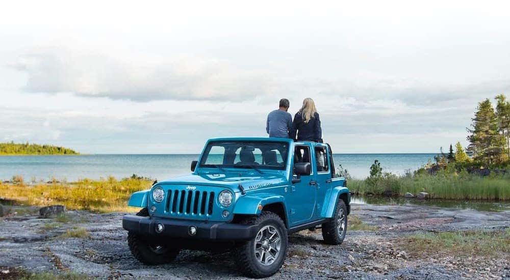 A blue 2021 Jeep Wrangler Rubicon is parked on a rocky shore next to a lake.