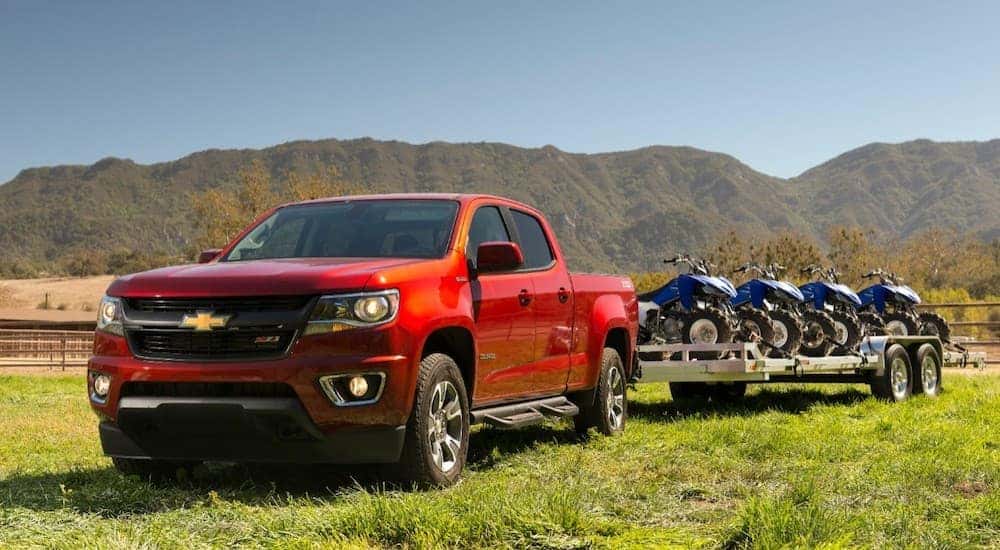 A red 2018 Chevy Colorado has four-wheelers on a trailer while on grass in front of mountains.