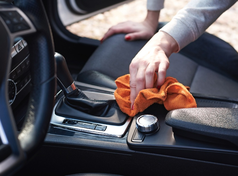 A hand with red nail polish uses an orange cloth to clean the console of a car interior. The image conveys tidiness and attention to detail.