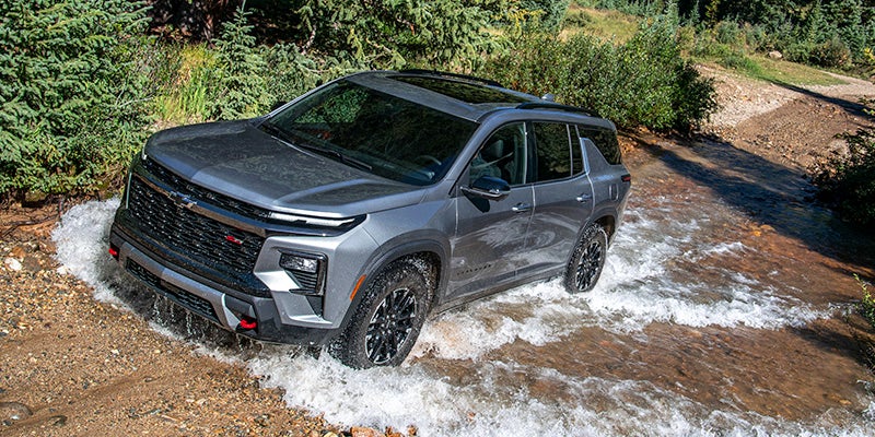 SUV driving through a shallow stream on an off-road trail, with water splashing around the wheels and a forested landscape in the background.