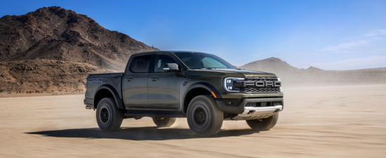 A rugged gray pickup truck drives across a sunlit desert landscape, kicking up dust. In the background, rocky hills rise under a clear blue sky, conveying adventure.