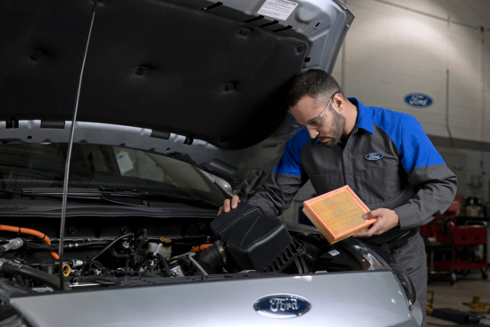 A mechanic in safety glasses and a uniform inspects a car engine under an open hood. He is holding an air filter. The setting is a Ford service center.