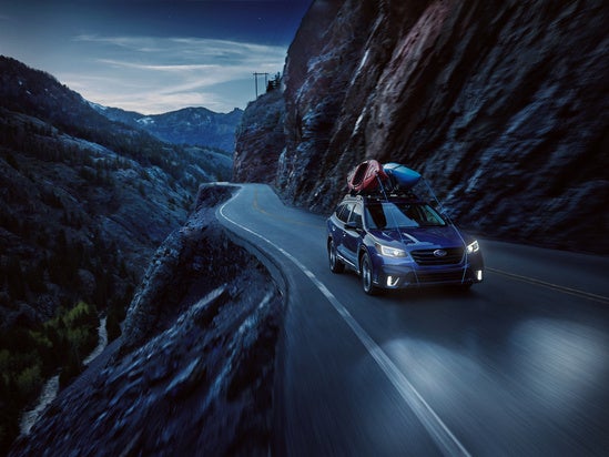 Subaru Outback driving on mountain road at night with its headlights on and gear on roof