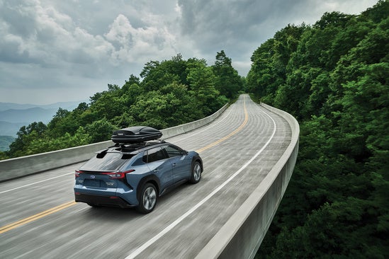 Subaru Solterra driving over a remote bridget with gear on its back surrounded by a forest canopy

