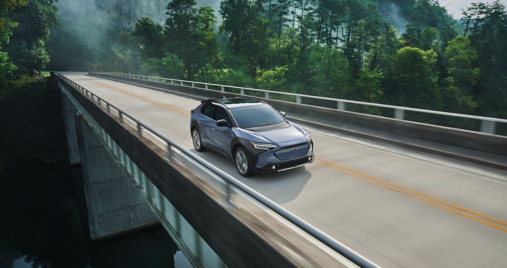 2026 Subaru Solterra driving across a bridge with lush green scenery in the background.