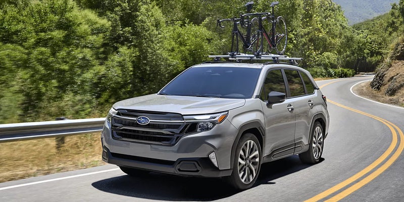 2026 Subaru Forester driving on a scenic mountain road with two bicycles mounted on the roof rack.