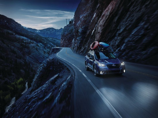 Subaru Outback with kayaks mounted to roof driving in mountains at night with headlights on